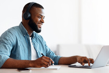 Poster - Online Education. Happy Black Guy In Headphones Study In Internet, Watching Webinar On Laptop Computer And Taking Notes, African Man Enjoying Distance Learning, Sitting At Desk At Home, Side View