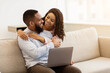 © Prostock-studio - Love And Tenderness. Beautiful african american couple relaxing together at home, using laptop, hugging. Loving wife emracing her husband from behind, man sitting on the sofa in living room or bedroom