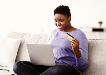 Poster - Online Shopping. Positive Smiling Young Black Girl Using Laptop And Credit Card At Home, Ordering Purchases In Internet And Making Payment, Sitting On The Couch In Living Room At Her Apartment