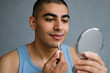 © smth.design - Young man in blue tank top applying lip gloss while looking in handheld mirror against neutral indoor background