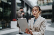 © qunica.com - A focused young woman in a beige blazer interacts with a large tablet in an outdoor professional setting. A blurred figure in the background adds depth to the scene.