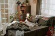 © AnnaStills - Bedroom featuring a messy bed with cozy pillows and blankets, surrounded by various plants and wooden furniture creating a warm and inviting atmosphere