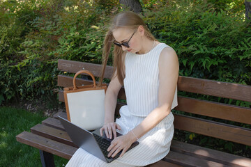 Poster - A woman is sitting on a bench and typing on a laptop. She is wearing sunglasses and a white dress