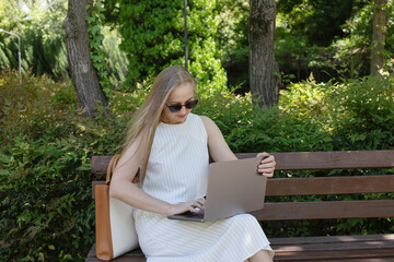Poster - A woman is sitting on a bench and typing on a laptop. She is wearing sunglasses and a white dress