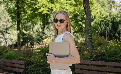 Poster - A woman is holding a laptop in a park. She is wearing sunglasses and a white dress. The scene is bright and sunny, and the woman is enjoying her time outdoors