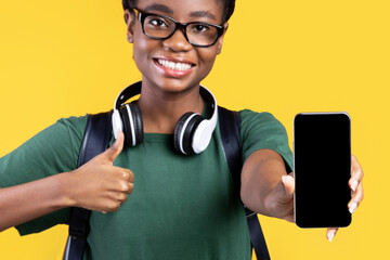Poster - Joyful Black Female Student Showing Smartphone With Empty Screen To Camera And Gesturing Thumbs Up Recommending Application For Cellphone Standing On Yellow Studio Background. I Like This App. Mockup