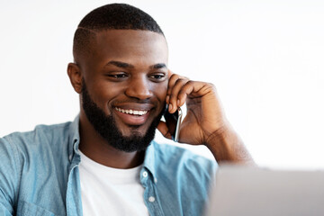 Poster - Closeup Portrait Of Smiling Black Man Talking On Cellphone And Using Laptop In Office. Handsome African American Entrepreneur Enjoying Phone Conversation While Working On Computer, Selective Focus