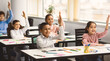 © Prostock-studio - Ready For An Answer. Diverse group of multicultural cheerful primary schoolkids sitting at table in classroom and raising hands, studying and learning. Portrait of positive little elementary students