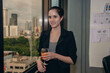 © S photographer - Professional woman holding a drink, dressed in a sleek outfit, with a city skyline backdrop, exuding confidence.
