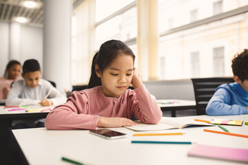Poster - Portrait of focused small asian girl sitting at table in classroom at primary school or kindergarten, reading task in notebook. Reopening and return back to school after coronavirus quarantine