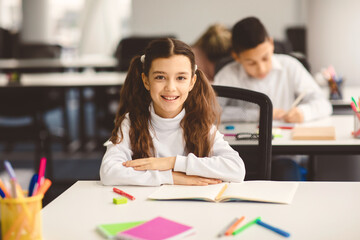 Poster - First Day At School. Portrait of smiling diligent small girl with ponytails sitting at table in classroom at elementary school with group of classmates in background and posing, looking at camera