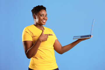 Poster - Cheerful African Woman Holding Laptop Pointing Finger At Computer Advertising Website Standing Over Blue Background, Smiling To Camera. Black Lady Posing With Laptop In Studio