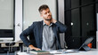 © Phushutter - A stressed Caucasian businessman in a suit sit indoors at an office desk, suffering from office syndrome symptoms including headache, neck pain, and wrist pain due to work-related strain.