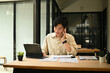 © saltdium - A Young Asian businessman talking on a mobile phone while working on a tablet at a modern office desk with financial documents
