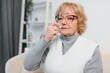 © Serhii - Blond senior woman sitting in chair at home and spraying nose spray