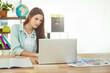 © wavebreak3 - Woman sitting at desk working on laptop with notebook, color printouts in home office, copy space