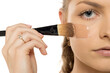 © vladimirfloyd - Close-up of woman applying liquid foundation with flat makeup brush on cheek, showing professional technique and smooth application against white background.