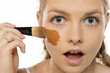 © vladimirfloyd - Close-up of surprised young woman applying foundation that's too dark for her skin tone, showing shocked expression while holding makeup brush against white background
