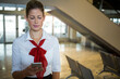 © Wavebreak Media - Female airline attendant holding smartphone and standing in gate area by escalator near metal seats