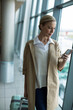 © Wavebreak Media - Woman standing holding smartphone at eye level beside rolling suitcase in airport terminal corridor