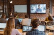 © andyaziz6 - A room of people sit at tables with laptops watching a presentation on a large screen at the front of the room