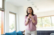 © Wavebreak Media - Adult woman holding microphone speaking in living room by sofa, blue cushions, glass door, window
