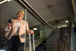 © Wavebreak Media - Mature female traveler in blazer holding phone and suitcase at airport escalator, copy space