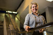 © Wavebreak Media - Woman in her fifties wearing shirt and cardigan smiling while holding booklet on lobby escalator