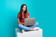 © deagreez - Young woman sitting with a laptop in a casual outfit against a teal background