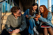 © Alberto - Three cheerful university students are sitting on campus stairs, sharing funny social media content on a smartphone and enjoying their time together