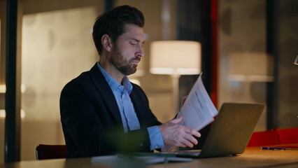 Poster - Late worker reading papers working at laptop in dark office closeup. Serious man