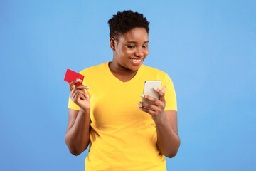 Poster - African Lady With Smartphone And Credit Card Using Mobile Banking Application Shopping Online Standing Over Blue Studio Background. Black Woman Using Phone App Concept