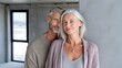 © AAS CREATIVE - A mature couple, he with short graying hair and she with shoulder-length gray hair, stand close, smiling subtly at the camera in an unfinished room, sunlight streaming in from a window behind them