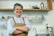 © Ekkasit A Siam - Asian senior female barista adult standing confidently with arms crossed smiling at camera inside cafe reflecting pride and success in small family coffee shop, Concept small business