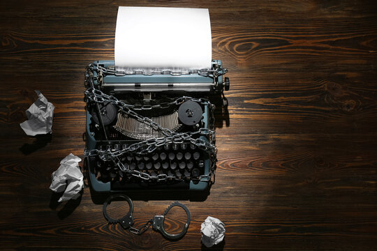 Vintage typewriter with chains, handcuffs and crumpled paper on wooden background. Printing ban concept