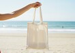 © Rehena - Hand holding a reusable tote bag on a sunny beach near the ocean ai generated