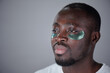 © pressmaster - Young Black person using green eye patches for reducing eye puffiness and improving skincare. Thoughtfully looking into distance, beads of sweat visible on skin