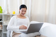 © Ekkasit A Siam - Asian pregnant woman sitting on sofa smiling using laptop wearing white sweater and brown pants resting hand on belly expressing joy relaxation preparing for future life with upcoming baby arrival