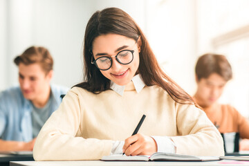 Poster - Studying And Learning Concept. Portrait of smiling young woman in eyeglasses sitting at table in lecture hall in highschool, writing in notebook, taking notes or examination, selective focus