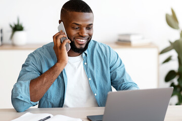 Poster - Remote Business. Smiling Black Male Freelancer Talking On Cellphone And Working On Laptop At Home Office, African Man Using Computer And Having Pleasant Phone Conversation, Enjoying Distance Job