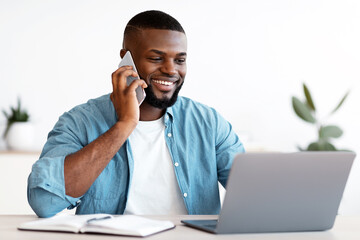 Poster - Black Male Entrepreneur Talking On Cellphone And Working On Laptop In Office. Smiling Millennial African American Businessman Sitting At Desk With Computer And Using Mobile Phone, Free Space