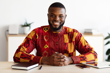 Poster - Portrait Of Handsome African Freelancer Man In Traditional Costume Sitting At Desk In Home Office, Positive Black Self-Employed Guy In Ethnic Costume Smiling And Looking At Camera, Free Space