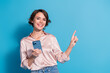 © deagreez - Smiling young woman in stylish satin blouse pointing at copy space against a blue background, holding smartphone