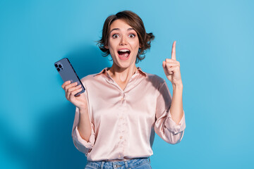 Poster - Young woman in pink blouse excitedly holding a smartphone with a surprised smile, pointing up against a vibrant blue background