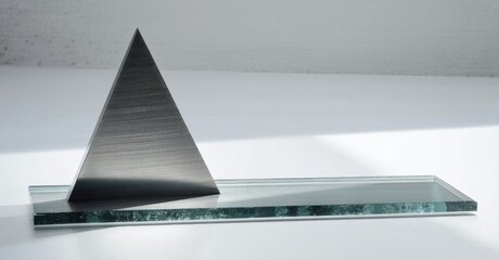 Poster - Dark pyramid rests upon rectangular glass plate. Minimalist composition, strong shadows, simple geometry, studio lighting. Clean aesthetic.