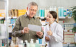 © JackF - In sales area of drugstore, mature man with prescription intently looks at display cases, trying to find vitamin for adults in open display. Girl pharmacist offer help to customer