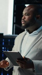 Poster - Vertical video IT professional looking over data center checkup checklist paperwork on tablet. African american man walking in server farm, reading documentation files on device, camera B