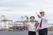 © kamonrat - A supervisor and secretary discuss logistics operations at a busy shipping port. Both wear safety helmets and hold documents, coordinating work amid cranes and stacked cargo containers.