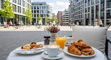 Outdoor coffee table in a lively city square with cappuccino, croissants, orange juice, and small pastries, surrounded by modern buildings and people walking