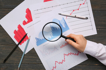 Fototapete - Female hand with magnifier, stationery and many business charts on brown wooden background